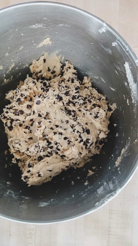 A close up of thick cookie dough in a mixing bowl with mini semi-sweet chocolate chips evenly distributed throughout the pale tan base.