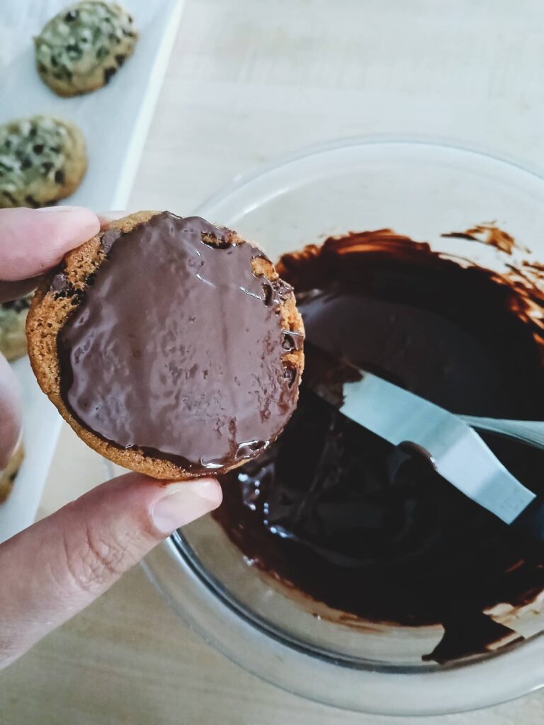 Using a small offset spatula to spread a thin, even layer of melted bittersweet chocolate onto the bottom of a cooled mini chip cookie.