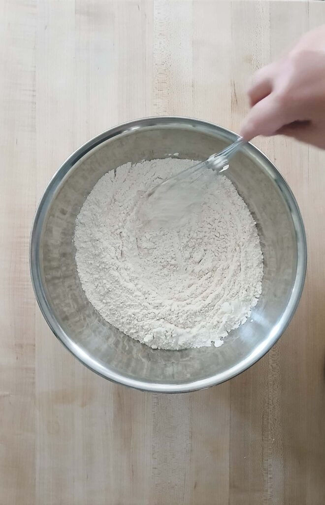 A hand whisking dry ingredients for cookie dough in a large metal bowl on a wooden countertop
