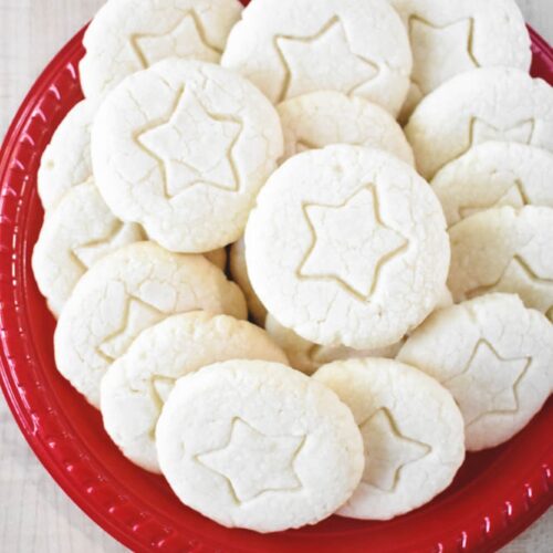 A red plate piled with round, pale whipped shortbread cookies featuring a delicate star imprint in the center of each.