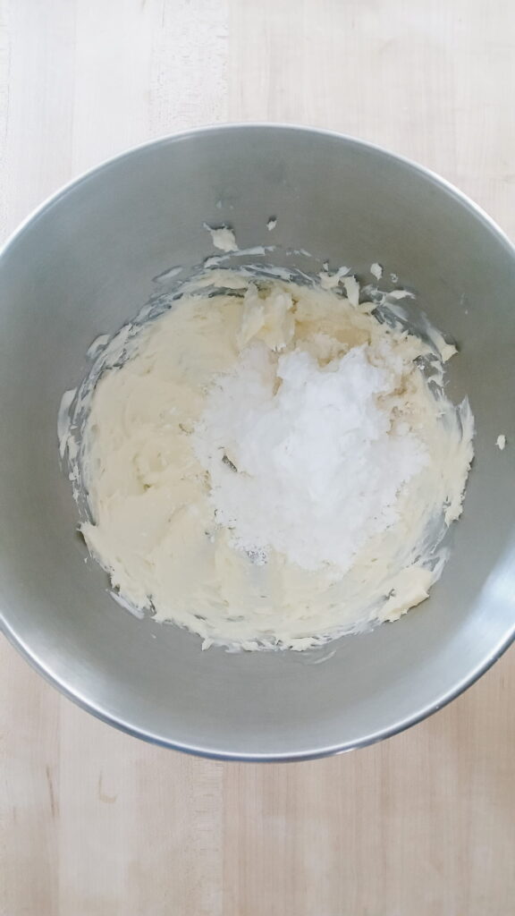 A stainless steel stand mixer bowl containing smooth butter topped with a mound of fine powdered sugar.