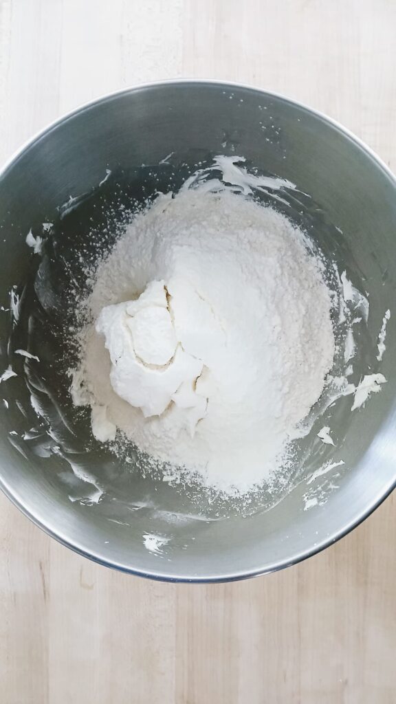 Adding all-purpose flour to the pale, whipped butter and sugar mixture in a metal stand mixer bowl for shortbread dough.